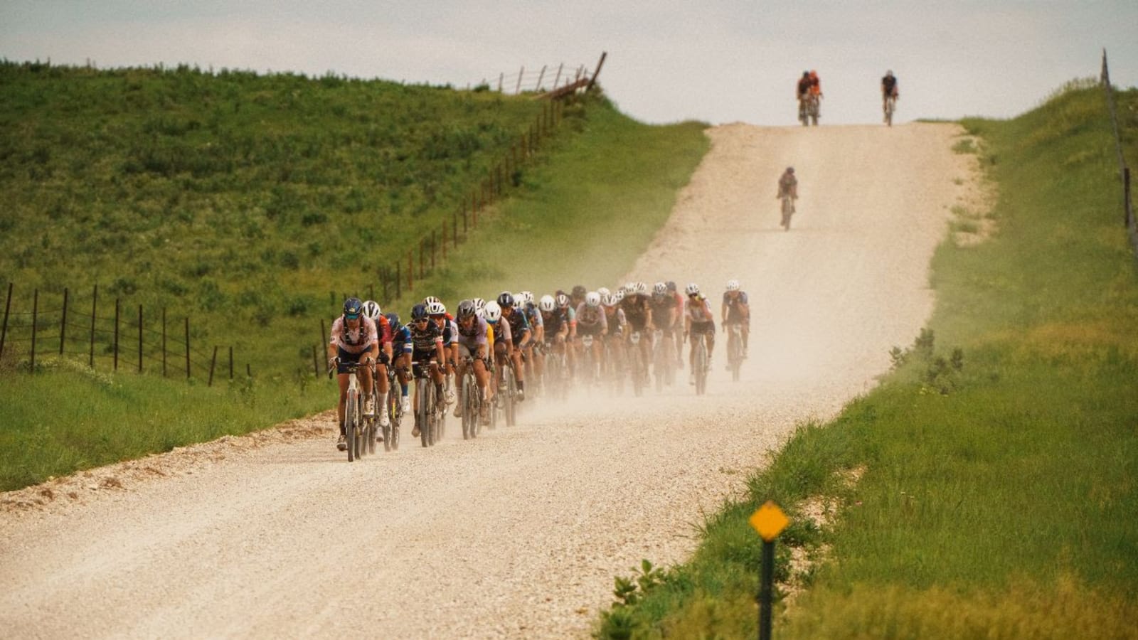 Pelotón de ciclistas de gravel rodando en fila por un camino de tierra entre campos